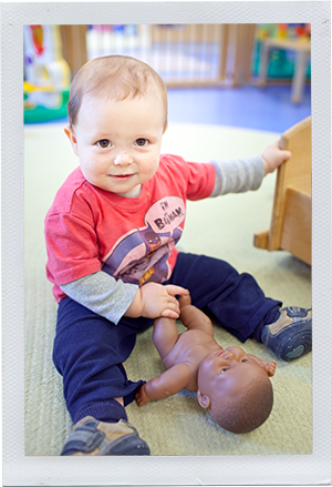 Photograph: An infant boy sits indoors smiling with a baby doll. (Photograph by Alex Lazara)