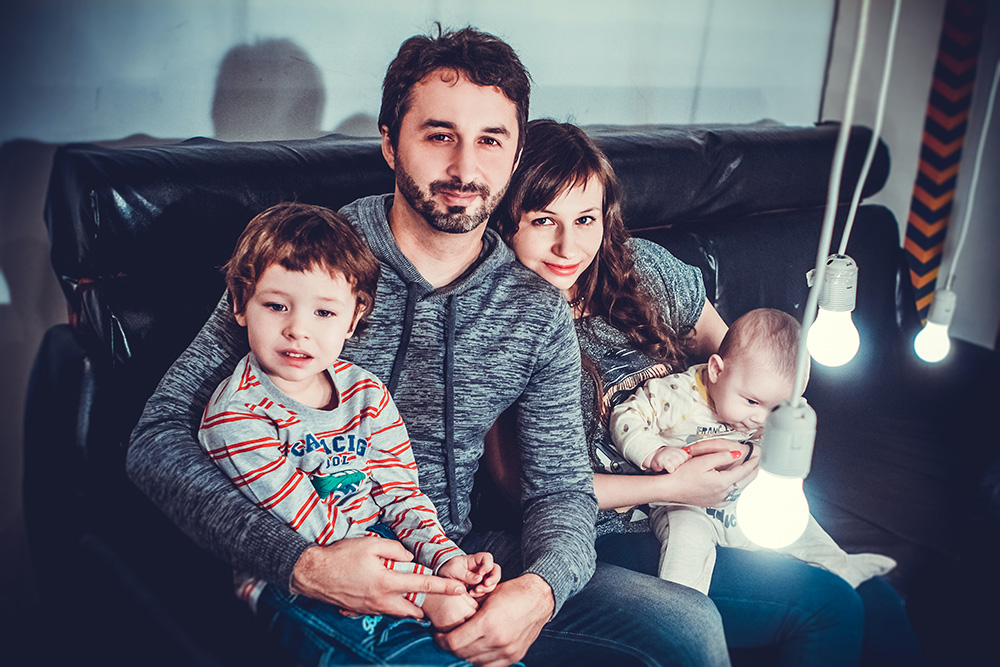 Photograph: a mother and father hold their two young children in thir laps. They are seated on a couch (Pexels, Victoria Borodinova).