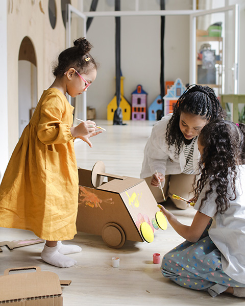 Photograph: A mother and her two children assemble and paint a cardboard car together.