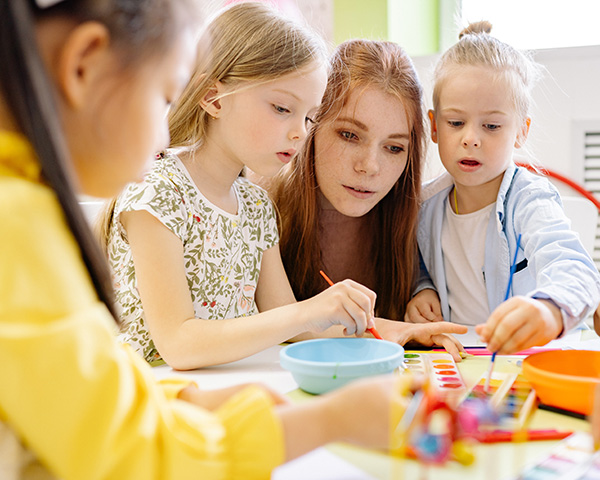 Photograph: A teacher looks on as three children paint at a classroom table.