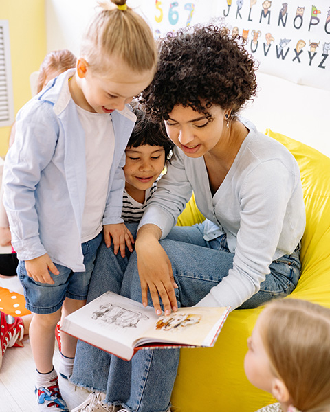 Photograph: A preschool teacher reads from a storybook. They are surrounded by four chldren.